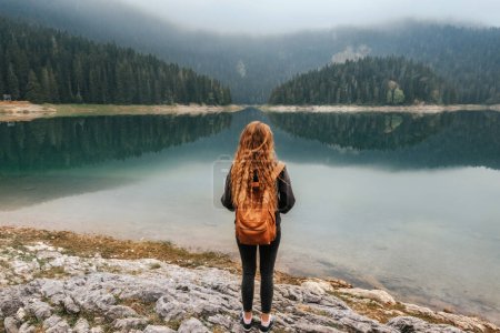 Foto de Mochilera mujer de pie sola en la orilla del lago de montaña y disfrutando de la vista del lago Negro en Durmitor. Viajera solitaria meditando disfrutando del silencio y del tiempo a solas, observando paisajes de lago brumoso. - Imagen libre de derechos