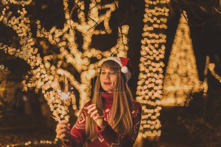 Photo for Happy woman in Santa hat and cozy winter sweater holding burning sparkler on background of golden Christmas tree lights. Smiling woman with dimples on cheeks celebrating the New Years Eve outdoors. - Royalty Free Image
