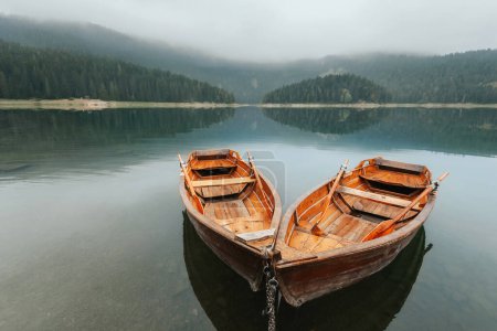 Foto de Dos botes de remos tradicionales en el lago de montaña cubiertos de niebla. Botes de remos en aguas tranquilas del lago Negro rodeado de bosque verde de coníferas en la mañana brumosa en el Parque Nacional Durmitor, Montenegro. - Imagen libre de derechos