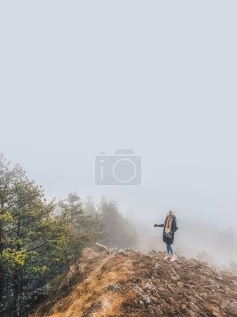 Photo for Woman standing alone on top of a rock in the middle of a foggy forest. Young female wearing winter clothes enjoying her walk in thick fog during winter holidays in mountains. - Royalty Free Image