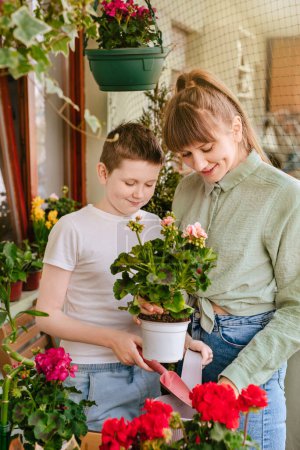 Photo for Happy mother and son spending time together in home garden. Cheerful boy and woman holding in hands pelargonium plant in pot, gardening at city balcony. Mom and kid working on urban terrace in spring. - Royalty Free Image