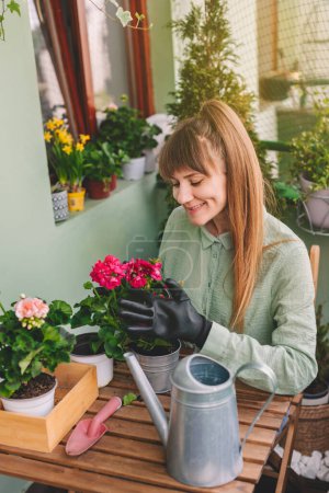 Photo for Happy woman gardener repotting flower sitting at wooden table in terrace garden. Young adult female in gardening gloves replanting geranium, using gardening tools. Home gardening at her balcony. - Royalty Free Image