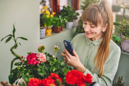 Woman gardener using mobile phone with gardening app for tips on caring for plants in terrace garden. Young female touching smartphone while caring plants of urban garden. Home gardening at balcony.