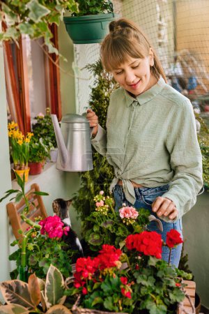 Photo for Happy woman gardener watering plants in terrace garden. Young adult female holding watering can taking care of blooming geranium and pelargonium flowers on green terrace. Home gardening at balcony. - Royalty Free Image