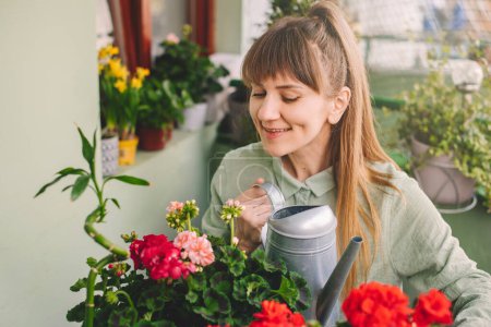 Photo for Happy woman gardener watering plants sitting at table in terrace garden. Young adult female holding watering can taking care of blooming geranium flowers on green terrace. Home gardening at balcony. - Royalty Free Image