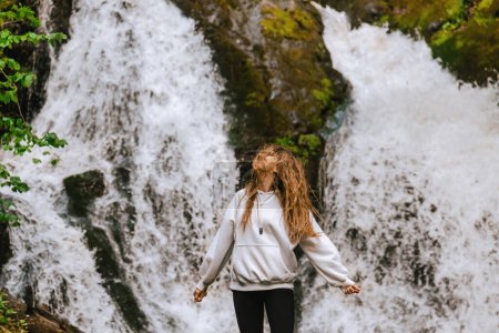 Mujer joven viajera con el pelo largo y volador disfrutando del paisaje de cascada de pie sola cerca de las cascadas de Izubra en el Parque Natural Golija, Serbia. Mujer viajando sola, armonía con el concepto de la naturaleza.