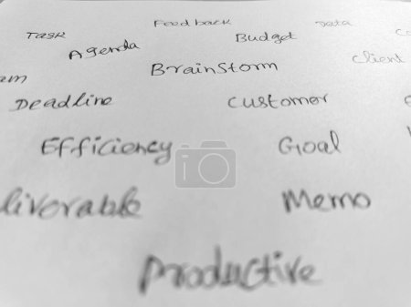 Close up of a white sheet of paper with various common office words handwritten in pen. black and white image. 