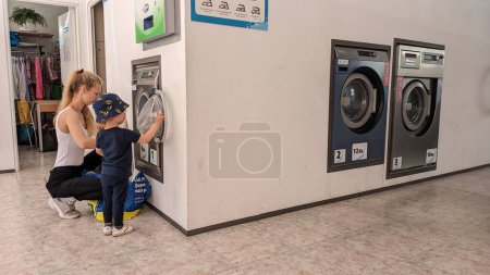 Téléchargez les photos : 17.03.20023 Jeune mère avec un jeune fils lavant ses vêtements dans une laverie automatique photo - en image libre de droit