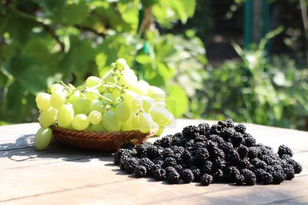 Close-up of fresh green grapes and black mulberries on a wooden table under sunlight in a garden setting.