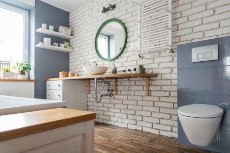 Téléchargez les photos : Intérieur moderne de salle de bain avec mur de briques blanches et meubles en bois et comptoir avec évier en céramique et toilettes élégantes. Décor design dans un loft industriel. - en image libre de droit