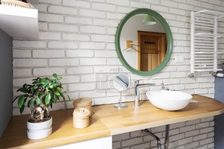 Foto de Interior industrial de baño con pared de ladrillo blanco, muebles de madera y mostrador con lavabo de cerámica y espejo redondo. Diseño moderno en apartamento loft. - Imagen libre de derechos