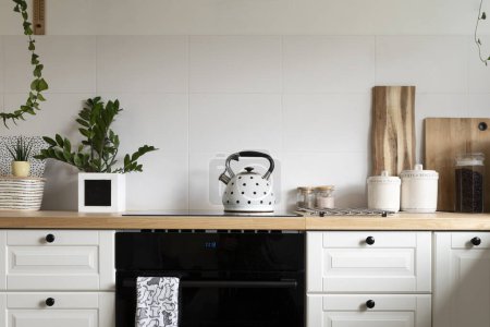 Photo for Modern interior of kitchen with wooden counter with kettle, white furniture with drawers and oven. Stylish room with kitchen utensils and scandinavian design at cozy home. White tiles on the wall. - Royalty Free Image