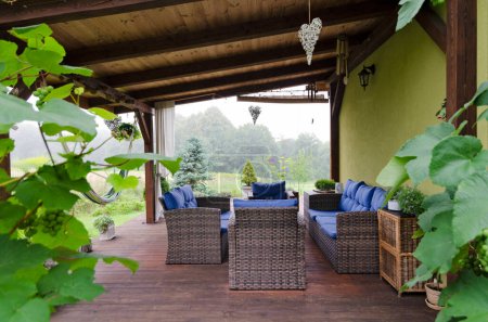 Beautiful covered terrace with rattan furniture table and armchairs, wooden floor and green leaves and plants. Outdoor design in garden on patio. Relaxation at veranda.