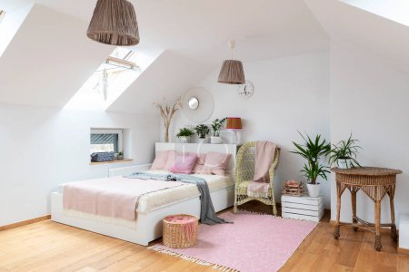 Photo for Cozy bedroom with doble bed with pink pillows and comfortable armchair in the attic. White wall with mirror, design lamp and carpet on the wooden floor in the room at home. - Royalty Free Image