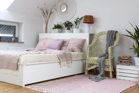 Téléchargez les photos : Chambre confortable dans le grenier avec mobilier blanc, lit avec oreillers roses et design, sol en bois et fauteuil. Intérieur lumineux dans un style scandinave. - en image libre de droit