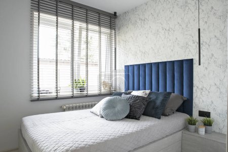 Foto de Cómoda cama con almohadas en el dormitorio con ventana con persianas. Cama en color azul marino y textura fondo de pantalla en la pared. Interior moderno en apartamento. - Imagen libre de derechos