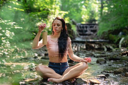 Téléchargez les photos : Jeune femme assise en position de yoga et pratiquer la thérapie sonore dans la forêt verte au milieu de la nature. Respiration, concentration et méditation avec sons. - en image libre de droit