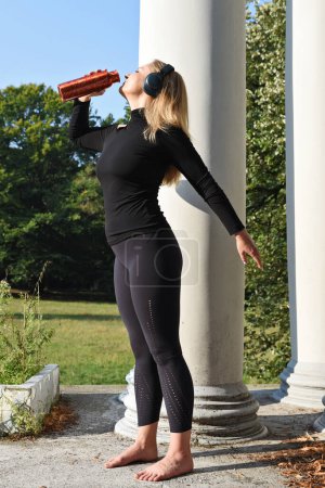 Foto de Mujer joven y activa en ropa deportiva negra después de entrenar y trotar con agua potable. Chica sedienta de pie bebe con botella de agua al aire libre.Vertical. - Imagen libre de derechos