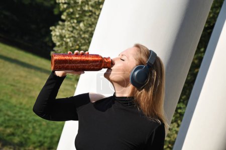 Téléchargez les photos : Portrait de jeune femme buvant de l'eau après s'être entraînée avec un casque dans un parc. Fille assoiffée avec bouteille d'eau pendant le sport extérieur. - en image libre de droit