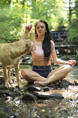 Téléchargez les photos : Femme de yoga assis dans la rivière avec chien et pratiquer la méditation et la relaxation. Mode de vie sain près de la nature dans la forêt. Auto-soin le matin. - en image libre de droit