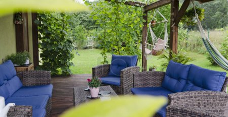 Téléchargez les photos : Terrasse couverte confortable avec fauteuils et canapé dans le jardin avec des feuilles vertes, de l'herbe et des fleurs. Lieu saisonnier pour la détente à l'extérieur de la maison. En plein air avec la nature.Bannière, panoramique. - en image libre de droit
