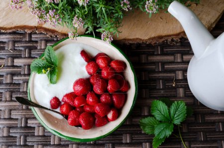 Photo for Ripe red strawberries with cream in a bowl on the wooden rustic table with green leaves and flowers. Healthy food, fruit for delicious dessert. Harvest in summer. Top view. Close up. - Royalty Free Image