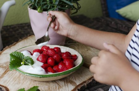 Photo for Little child holds in a hand spoon with strawberry. Red ripe strawberries in a bowl for healthy dessert. Food in a garden from summer hanvest on a wooden rustic table. Close up. - Royalty Free Image