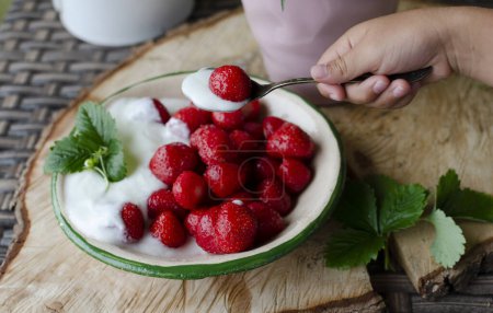 Photo for Little child holds in a hand spoon with strawberry. Red ripe strawberries in a bowl for healthy dessert. Food in a garden from summer hanvest on a wooden rustic table. Close up. - Royalty Free Image