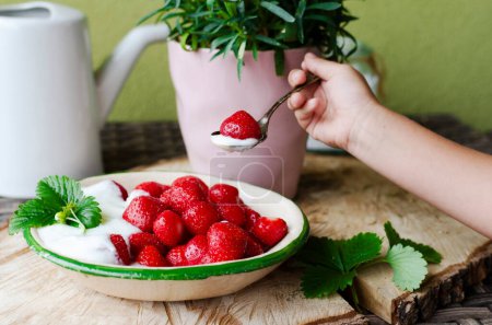 Photo for Spoon with strawberry in child hand and red ripe fruits with sweet cream in the bowl. Ecological natural strawberries as a healthy food. Close up. - Royalty Free Image