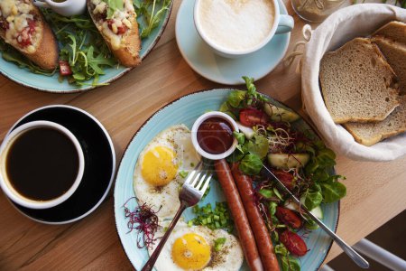 Photo for Plate with tasty food for brakfast with cup of coffee on a table in restaurant. Fried eggs, sausage and vegetable salad as a helthy meal. Different colors of food for eat. - Royalty Free Image