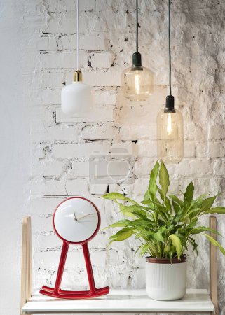 Foto de Las lámparas de diseño iluminan un estante en la oficina con un moderno reloj rojo y una planta sobre el fondo de ladrillos blancos. Vista vertical. - Imagen libre de derechos