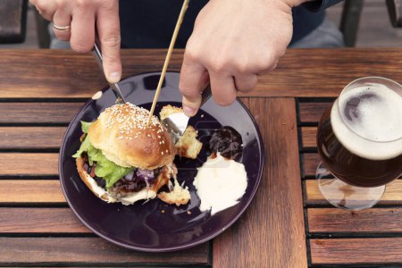 Téléchargez les photos : Burger sur une assiette et les mains avec couteau couper le hamburger. Restauration rapide saine au restaurant avec des légumes et de la bière. - en image libre de droit