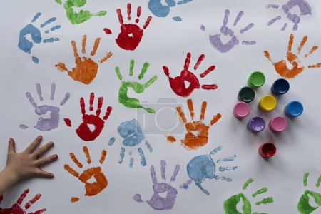 Téléchargez les photos : Empreintes de mains colorées sur papier blanc, peintures et main de petit enfant. Arrière-plan avec des empreintes dans des couleurs différentes. Éducation par le jeu, le plaisir et la créativité. Sensoriel à la maternelle. - en image libre de droit