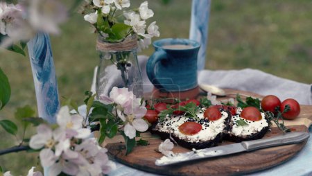 Photo for Rural breakfast in the garden on chair with blooming flowers and homemade slice of bread and tomatoes and mug of coffee in blue color. Healthy natural food outdoors. Close up. - Royalty Free Image