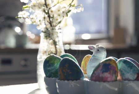 Photo for Homemade painted easter eggs with easter bunny on a table in the kitchen with sun rays from the window and white spring flowers. Holiday time and breakfast celebration. Close up. - Royalty Free Image