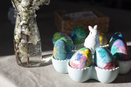 Téléchargez les photos : Œufs de Pâques colorés avec lapin de Pâques sur une table avec des fleurs blanches dans un vase en verre. Printemps et décoration. Lumière dans la cuisine. Horizontal. - en image libre de droit