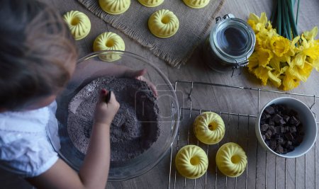 Téléchargez les photos : Mains de petit enfant avec cuillère mélanger une pâtisserie pour des muffins faits maison avec du chocolat. Chef dans la cuisine préparer le dessert pour Pâques à la maison. Forme pour la cuisson des cupcakes. Vue du dessus. - en image libre de droit