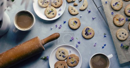 Homemade cookies with blue edible flowers with cups of coffee and rolling pin on blue background. Table full of healthy dessert for vegan. Food and crunchy biscuit.