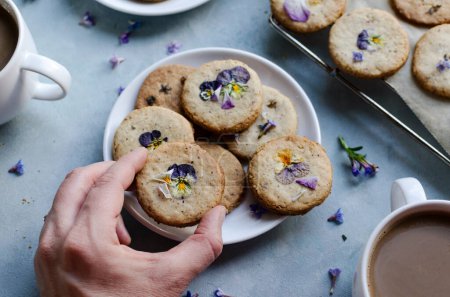 Hand woman with homade cookie with edible flowers. Dessert in spring time and healthy snack on a plate with cup of coffee. Freshly baked on blue background. Flat lay food. Close up.