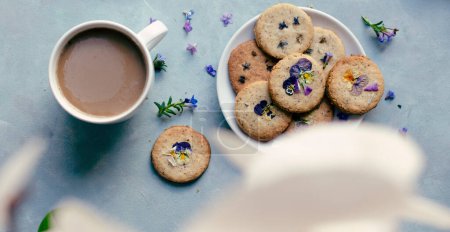 Cookies with edible flowers on a plate with cup of coffee on a blue background in vintage style. Homemade biscuit with spring flowers. Decorated food. Top view. Panoramic.