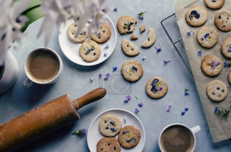 Homemade cookies with blue edible flowers with cups of coffee and rolling pin on blue background. Table full of healthy dessert for vegan. Flat lay food and crunchy biscuit. Top view.