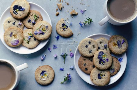 Food on a table for healthy dessert. Buscuits on a plates with edible flowers with cups of coffee on blue background. Homemade cookies in spring time. Dessert. Top view.