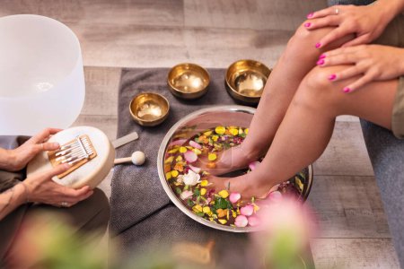 Female feet at spa salon with sound healing. Legs of woman in bowl with water decoration flowers and petals of rose. Massage and pedicure. Relax.
