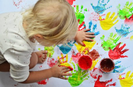 Téléchargez les photos : Petite fille peint à l'école maternelle sur fond blanc. Empreinte multicolore des mains et plaisir de l'enfant. - en image libre de droit