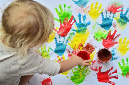 Little girl paints in nursey school on white background. Multicolor imprint of hands and fun of child.