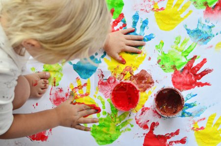 Little girl plays multicolor paints and imprints hands.