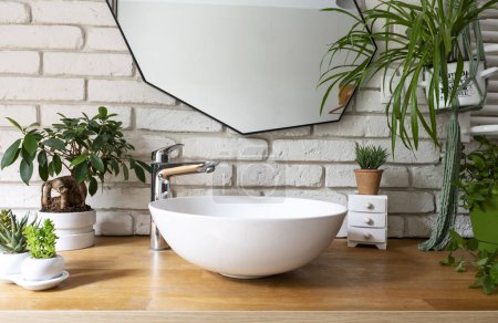 Photo for Modern interior with mirror on white brick wall and stylish sink and faucet in industrial bathroom. Bright bath with green plants and wooden furniture. - Royalty Free Image