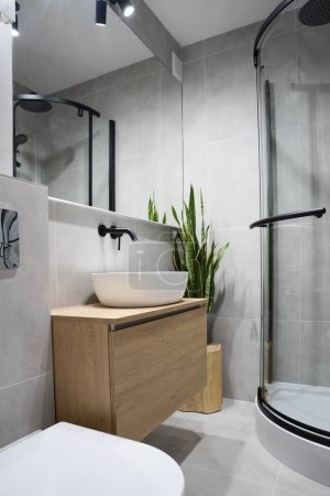 Foto de Cuarto de baño moderno con azulejos grises, muebles de madera, ducha, lavabo elegante y espejo. Interior industrial en casa. Vertical. - Imagen libre de derechos