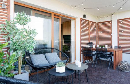 Téléchargez les photos : Belle terrasse avec du bois sur le mur, table en bois et canapé confortable. Patio avec beaucoup de plantes et grande fenêtre. Espace confortable extérieur. - en image libre de droit