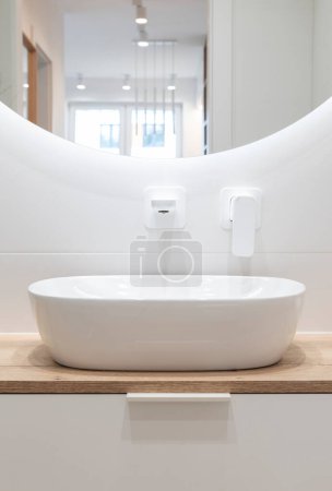 Téléchargez les photos : Salle de bain moderne avec carrelage blanc, lavabo en céramique et robinet sur comptoir en bois avec armoire. Design luxueux et élégant dans le nouvel intérieur de l'appartement. - en image libre de droit